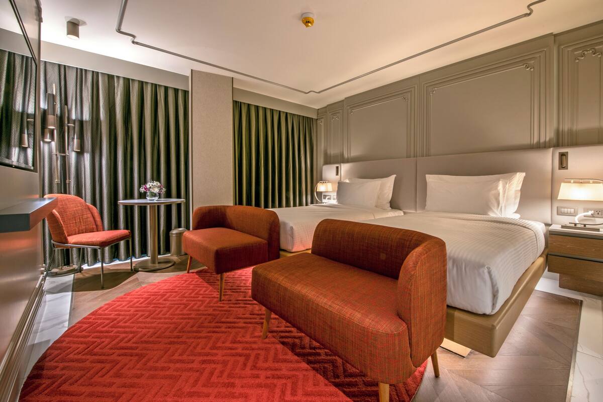 A hotel room with a large bed, two armchairs, a small table with a vase, and a red carpet.