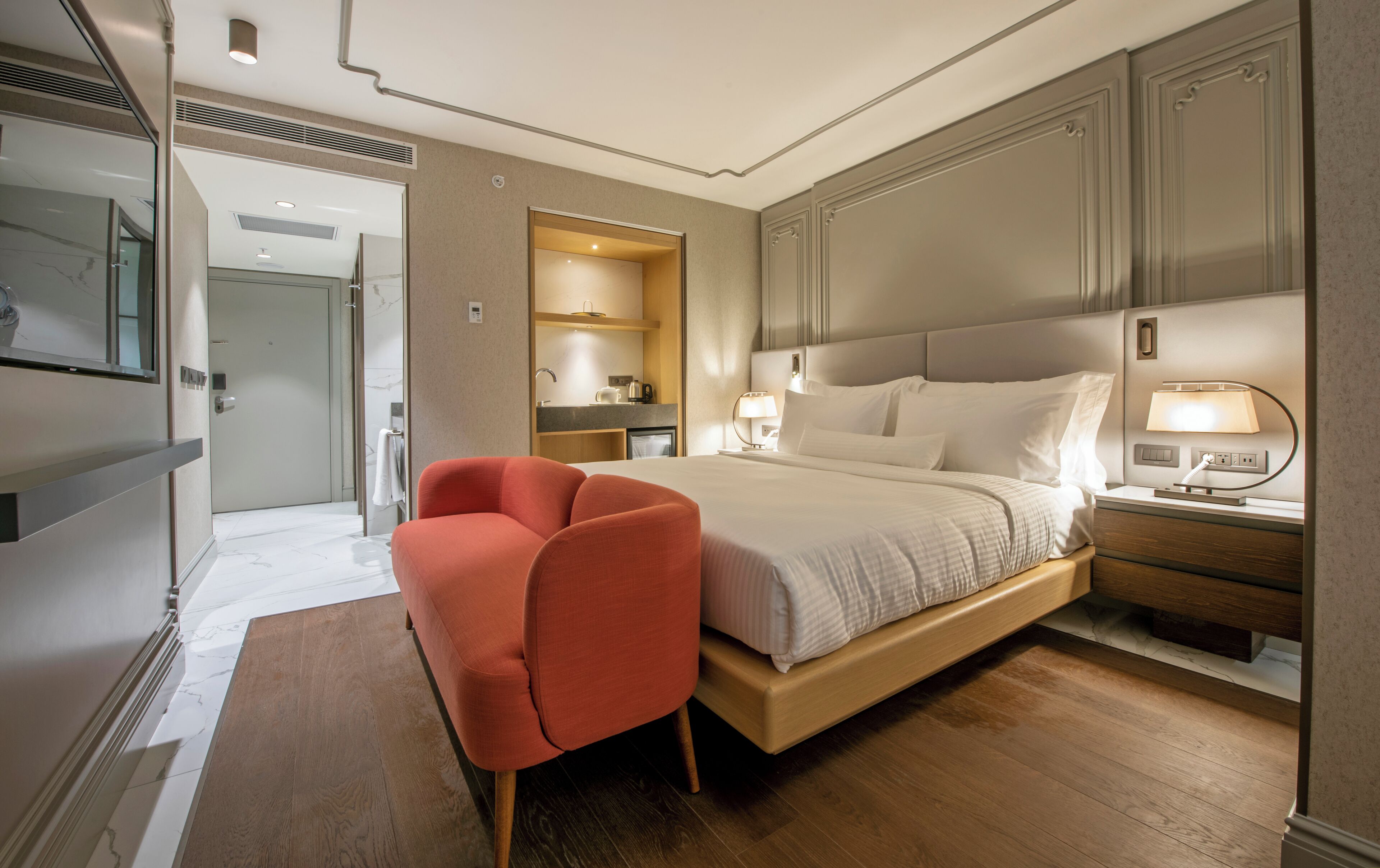 A modern hotel room with a large bed, a sitting area with a red chair, a bedside table with a lamp, and a bathroom visible through an open door.