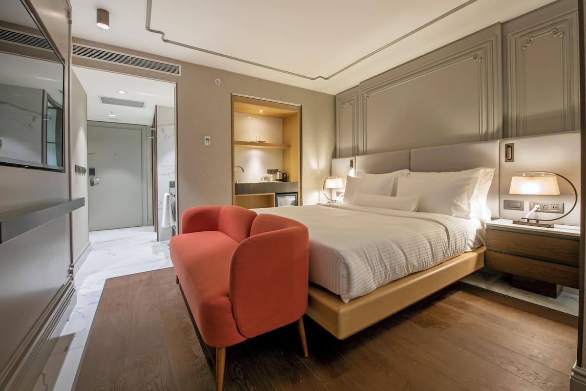 A modern hotel room with a large bed, a sitting area with a red chair, a bedside table with a lamp, and a bathroom visible through an open door.