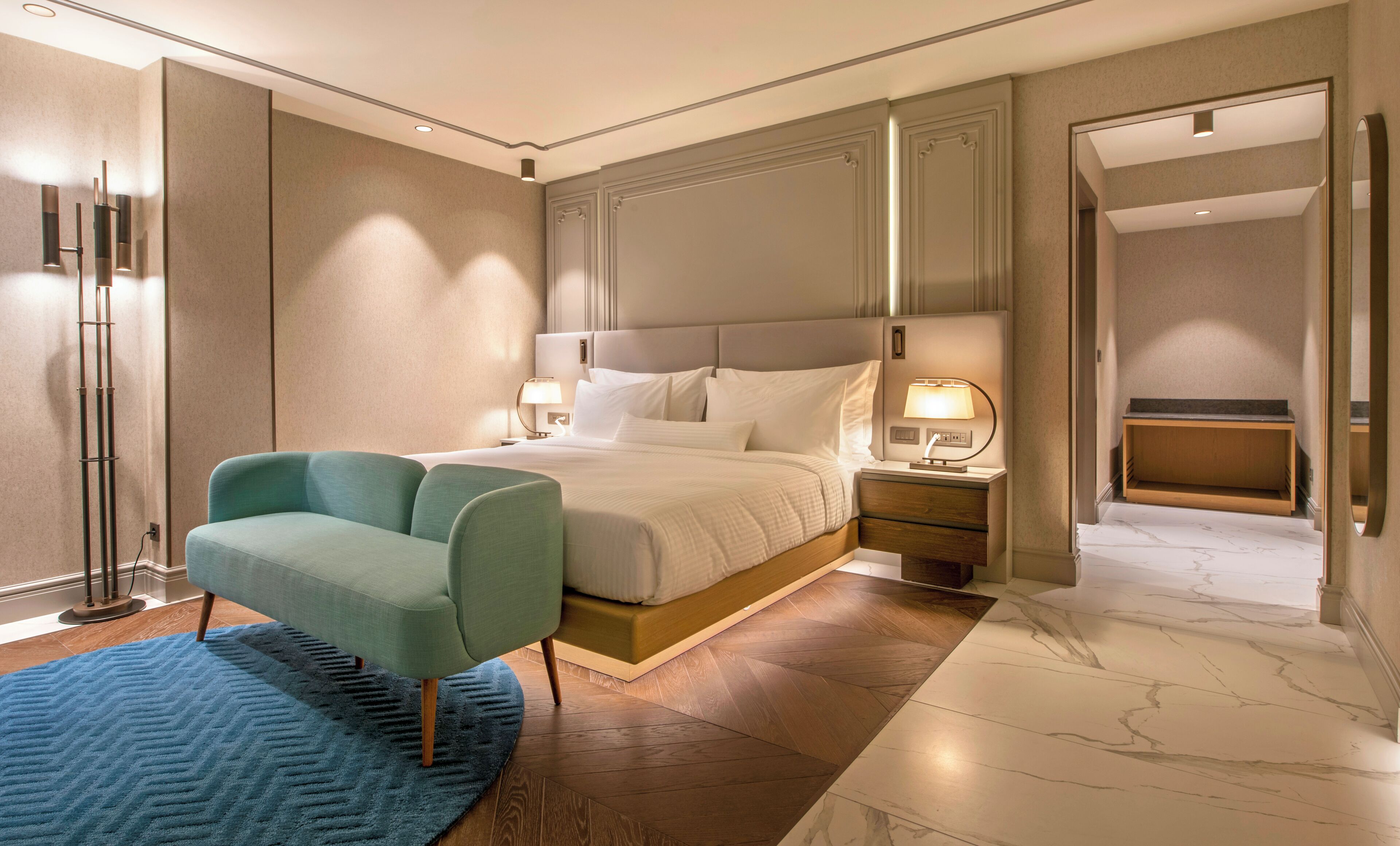 A modern hotel room with a large bed, a seating area, a nightstand with a lamp, and a bathroom visible through an open door.