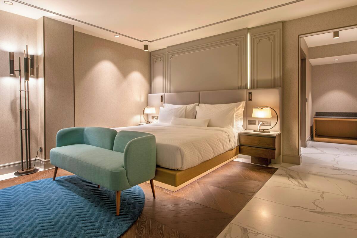 A modern hotel room with a large bed, a seating area, a nightstand with a lamp, and a bathroom visible through an open door.
