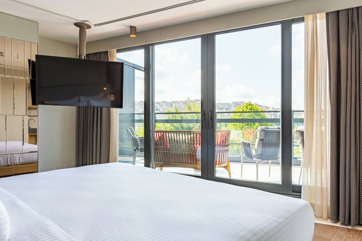 A hotel room with a large bed, a TV mounted on the wall, and a balcony with outdoor seating.