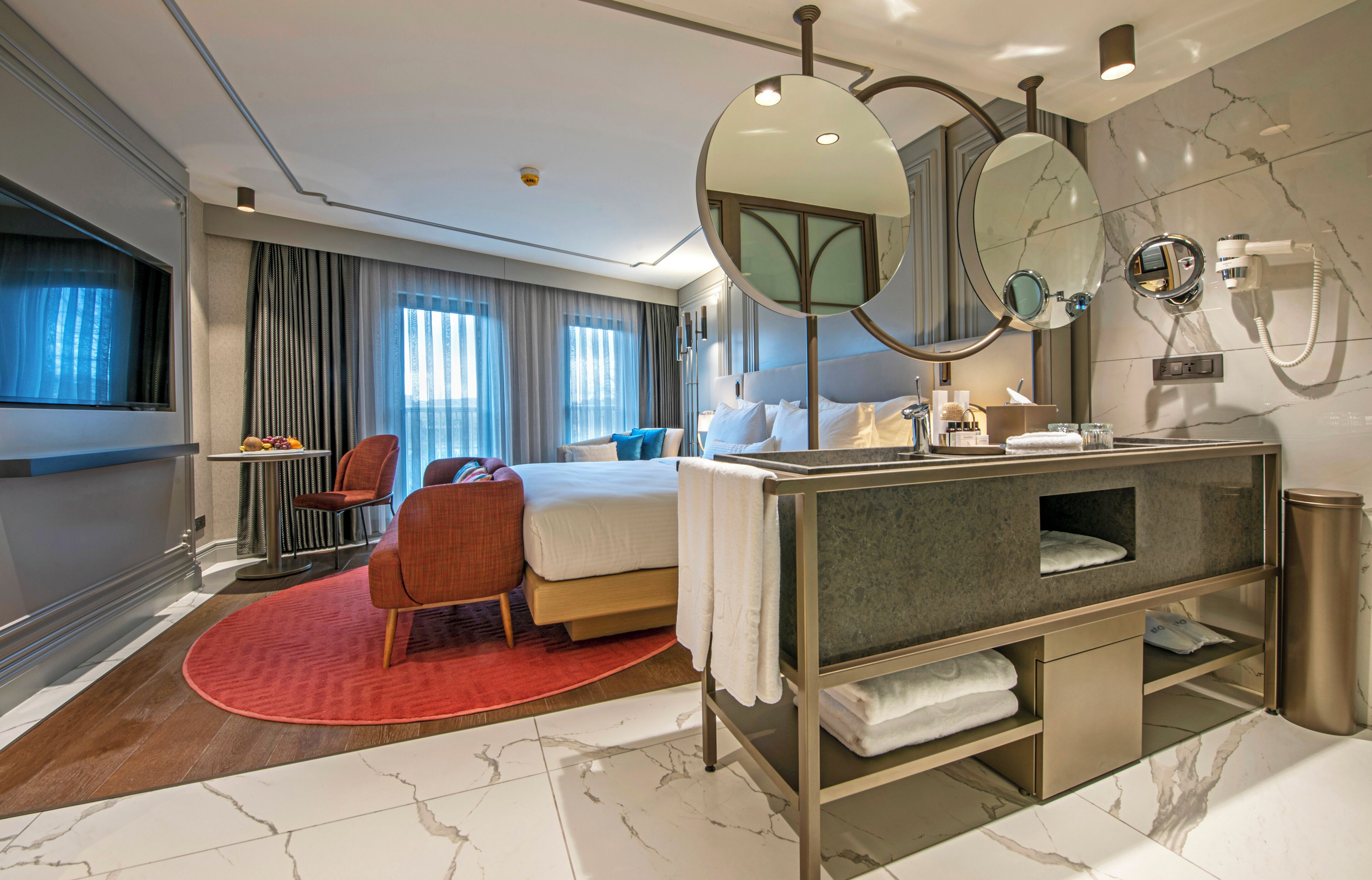 A modern hotel room with a bed, a seating area with a chair, a vanity with a mirror, and a bathroom with a toilet and sink.