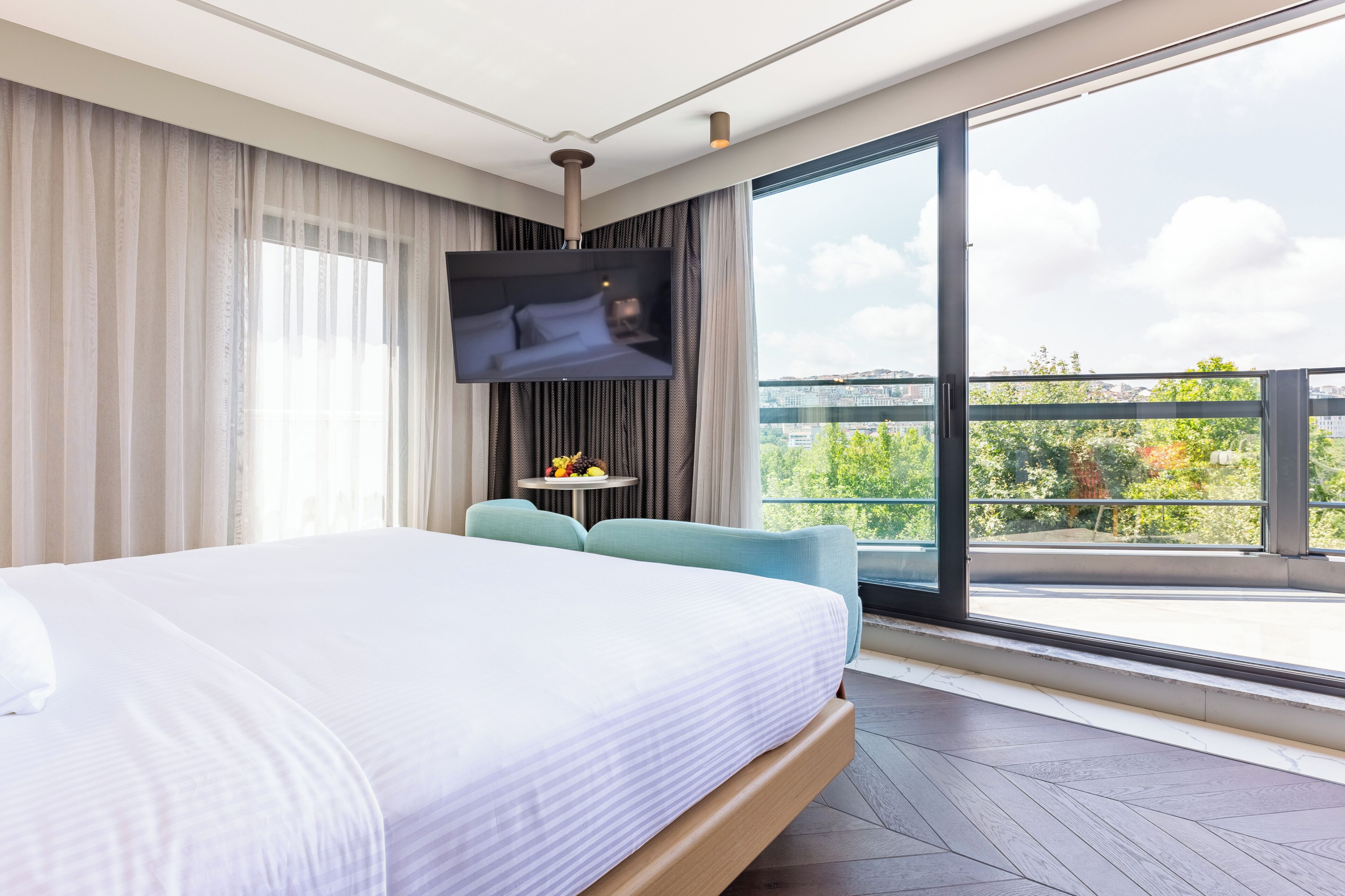 A modern hotel room with a large bed, a TV mounted on the wall, a small table with a fruit bowl, and a balcony with a city view.