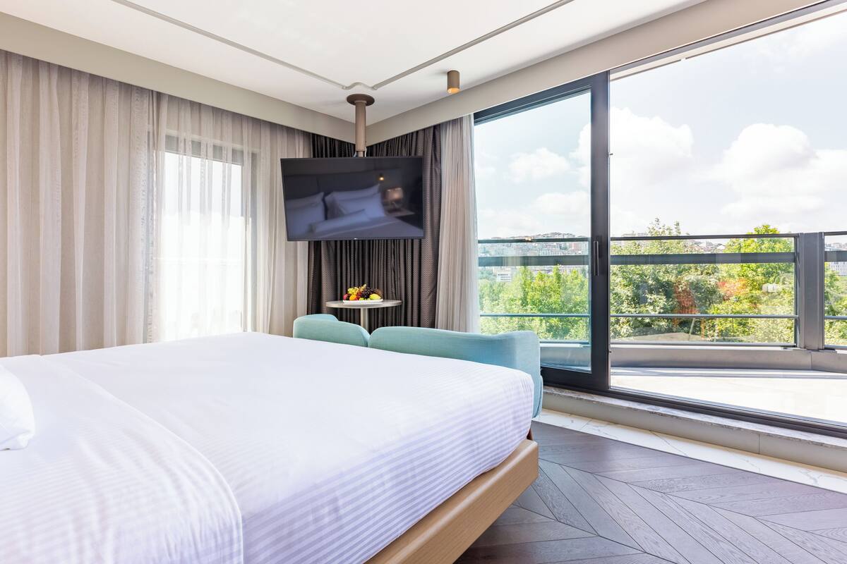 A modern hotel room with a large bed, a TV mounted on the wall, a small table with a fruit bowl, and a balcony with a city view.