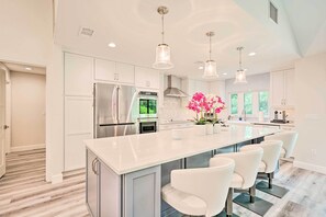 Une cuisine moderne avec des meubles blancs, des appareils en acier inoxydable, un grand îlot central doté de tabourets et un éclairage suspendu.