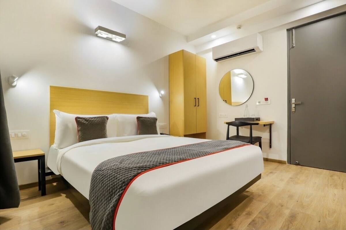 A modern hotel room with a large bed, a bedside table, a small desk, a mirror, and a wardrobe.