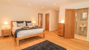 A bedroom with a wooden bed, two bedside tables with lamps, a dark rug, and a bathroom with a sink and mirror. - Sardis Chapel 1 (Bodorgan)