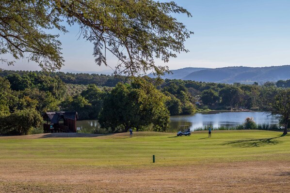 Un campo de golf con un club de campo, un lago y colinas al fondo.