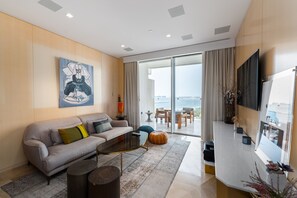 Een moderne woonkamer met een grijze bank, een salontafel en een grote televisie. Er is een schuifdeur naar een balkon met uitzicht op de stad.