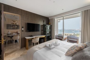 A modern hotel room with a large bed, a desk, a TV, and a view of the sea.