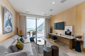 A modern living room with a sofa, coffee table, and TV. It features a large window with a view of buildings and a balcony.