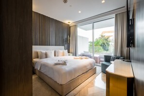 A modern hotel room with a large bed, a flat-screen TV, a sofa, and a large window offering a view of the outdoors.