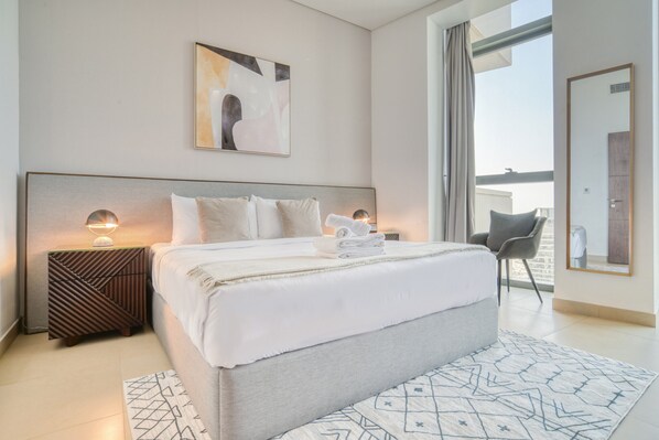 Une chambre d’hôtel moderne avec un grand lit, une table de chevet avec une lampe, une chaise et une vue sur l’extérieur.