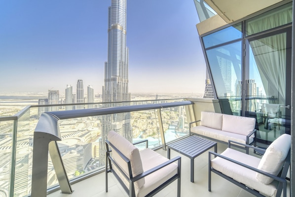 A balcony with a city view, including a tall skyscraper, furnished seating, and a glass railing.