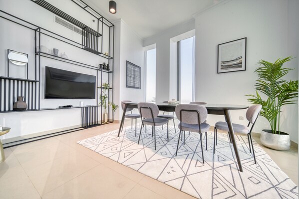 A modern living room with a flat-screen TV, a dining table with chairs, a large potted plant, and a geometric patterned rug.