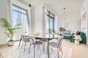 A modern living room with a round glass table, grey chairs, a green sofa, and a large window offering a city view.