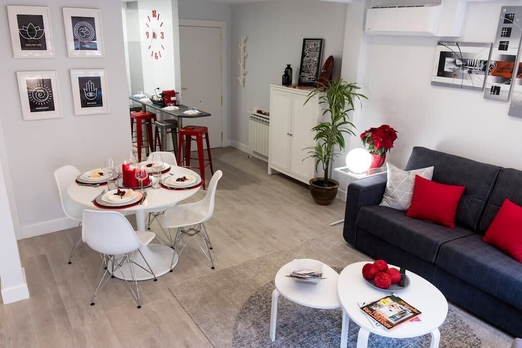Un salon moderne avec un canapé gris foncé, des coussins rouges et une table basse blanche. On y trouve également un coin repas avec une table et des chaises blanches, et une cuisine en arrière-plan.