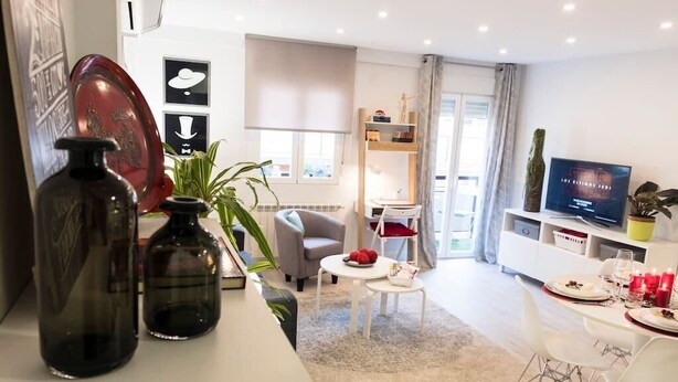 Un salon moderne avec un canapé, une table basse et un coin repas. On y trouve également un téléviseur, une bibliothèque et une plante.