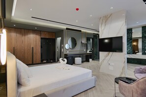 A modern hotel room with a large bed, a TV, a vanity with a mirror, and a seating area with a chair and a small table. - Adamas Resort & Hotel (Shënkoll)