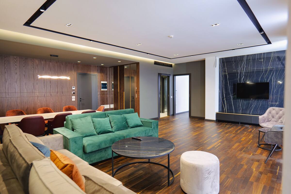 A modern hotel lobby with a wooden panel wall, a large seating area with sofas and chairs, a round table, and a flat-screen TV mounted on the wall.