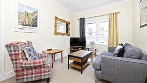 A living room with a plaid armchair, a grey sofa, a wooden coffee table, a flat-screen TV, and a framed picture of Glen Ogle. - Modern Flat on Rose Street (Edinburgh)