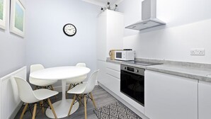 A modern kitchen with white cabinetry, a round dining table with chairs, a microwave, an oven, and a clock on the wall. - Modern Flat on Rose Street (Edinburgh)
