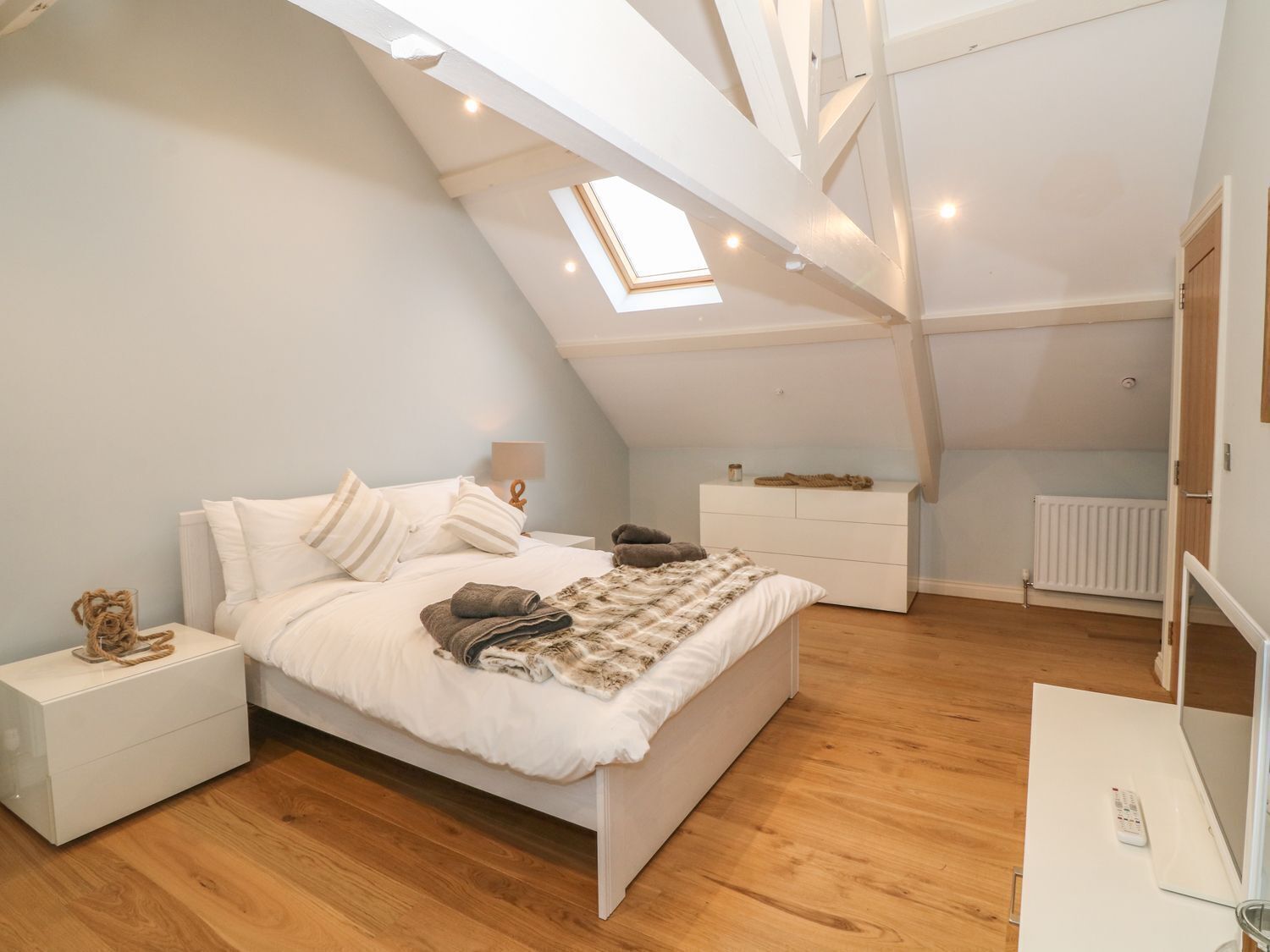 A modern, well-lit attic bedroom with a sloped ceiling, a skylight, a bed with white linens, a bedside table, a dresser, and a mirror.