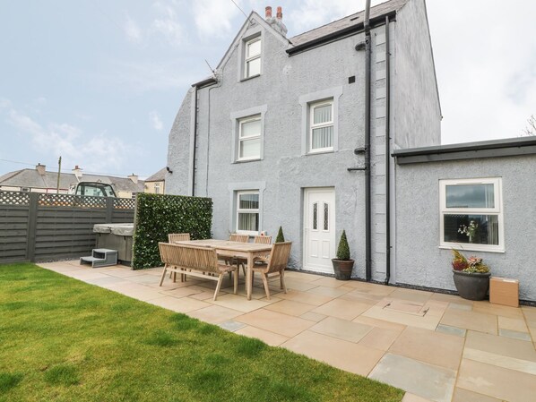 A two-story house with a patio area featuring a wooden table and chairs, a small garden, and a grey exterior wall. - Fishermans Cottage (Bodorgan)