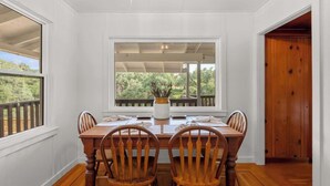 A dining area with a wooden table and chairs, white walls, and a view of greenery outside.