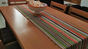Un coin repas avec une table en bois, des chaises, un panier de pain, une nappe à rayures, un téléviseur accroché au mur et une fenêtre avec des stores.