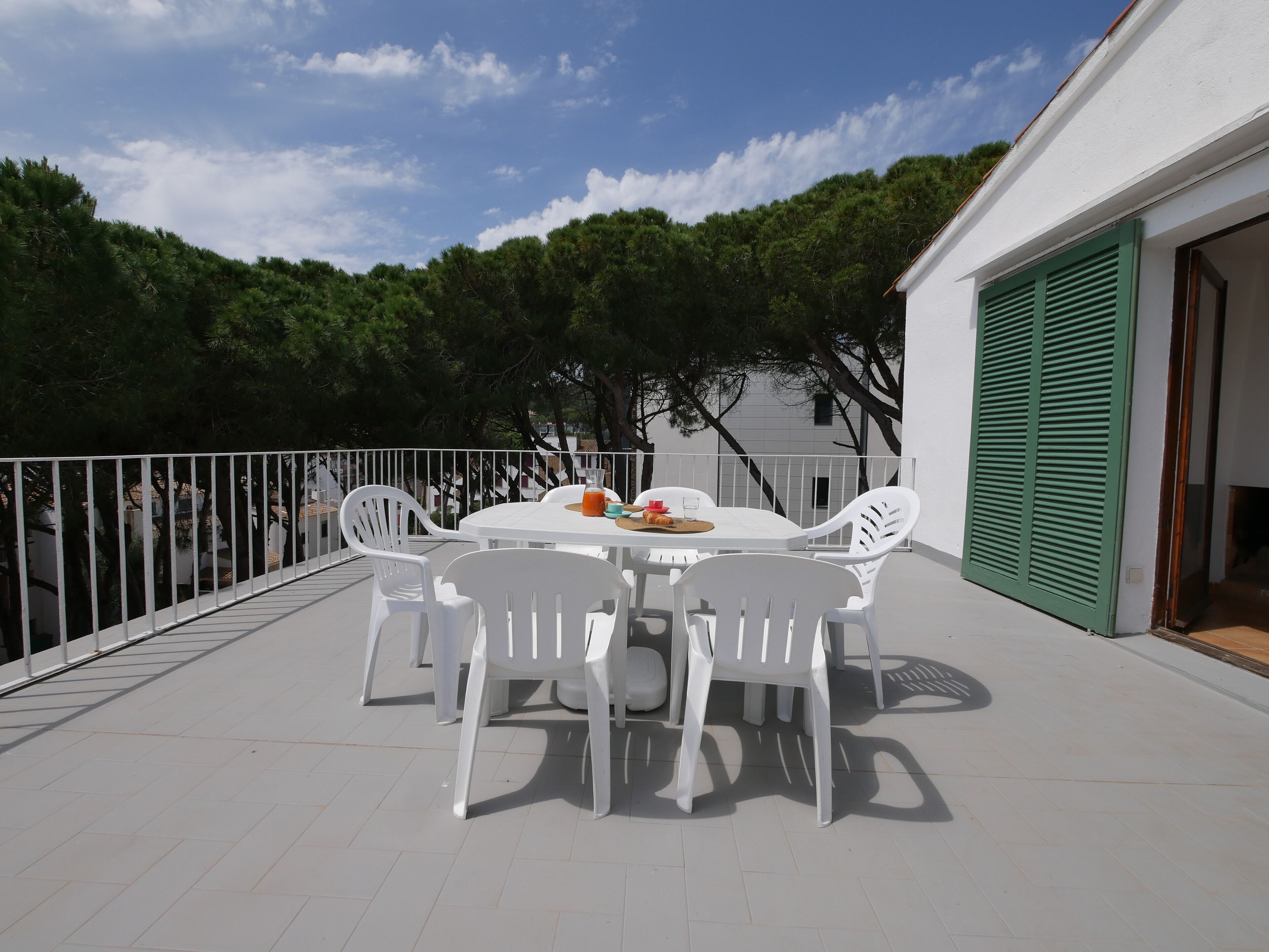 Un patio esterno bianco con un tavolo rotondo e quattro sedie bianche, una porta verde con persiane e un parapetto bianco.