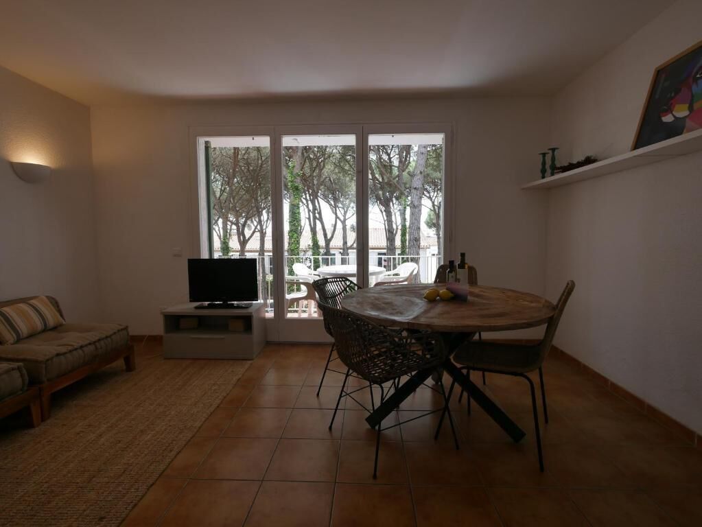 Una sala de estar con sofá, televisor, mesa de comedor con sillas y un amplio ventanal con vista a árboles y un balcón.
