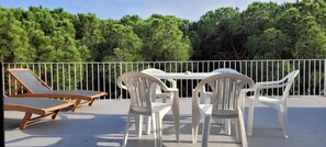 Une terrasse avec des chaises et une table en plastique blanc, entourée d’une clôture en métal blanc, et en arrière-plan, des arbres verdoyants.