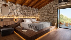 Un dormitorio con pared de piedra, vigas de madera en el techo, una cama con manta estampada, mesitas de noche y vistas al exterior.