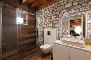 A bathroom with a stone wall, wooden floor, a glass shower enclosure, a toilet, and a sink with a mirror.