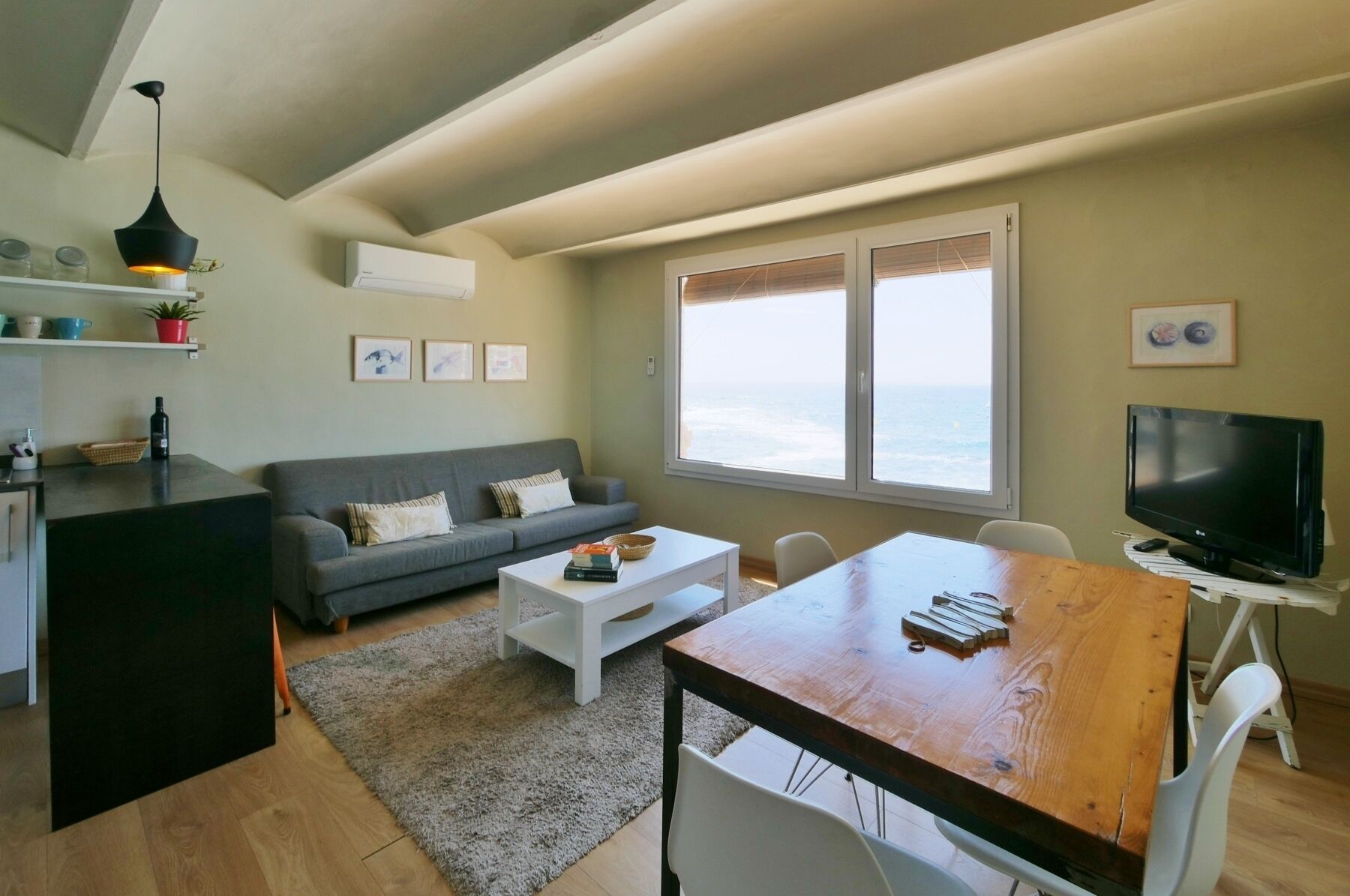 Ein Wohnzimmer mit grauem Sofa, einem Holzesstisch, einem Fernseher und einem großen Fenster mit Meerblick.