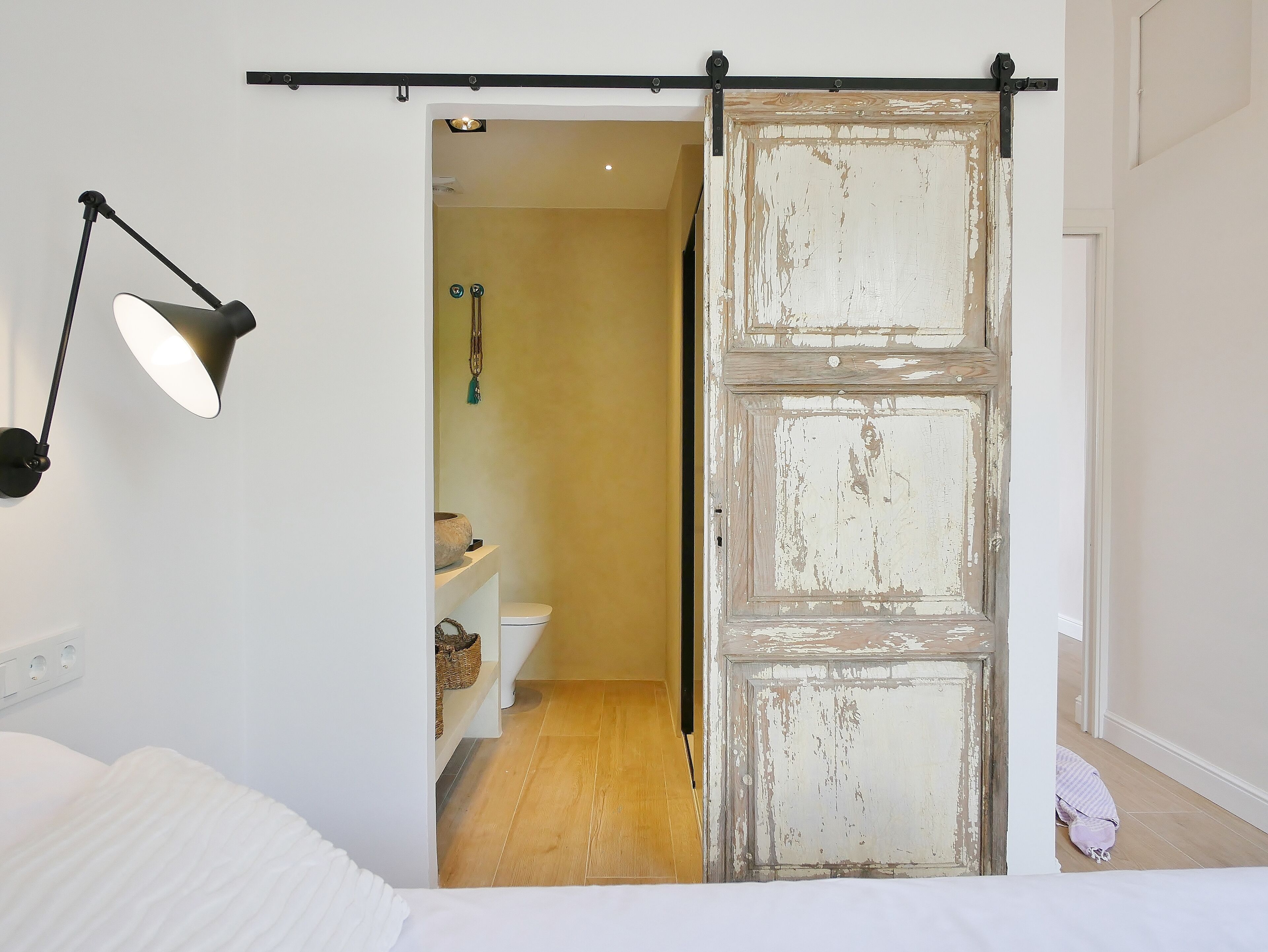 A bedroom with a bed, a bedside lamp, a sliding door leading to a bathroom, and a small shelf with a bowl and a hanging ornament.