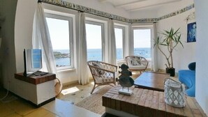 A room with a wooden coffee table, two wicker chairs, a small statue, and a view of the sea through large windows. -  Casa Concha Apartamentos (Palafrugell)
