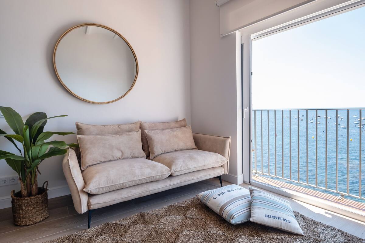 Una sala de estar moderna con un sofá color beige, un espejo redondo, un ventanal con vistas al mar y una planta en maceta.