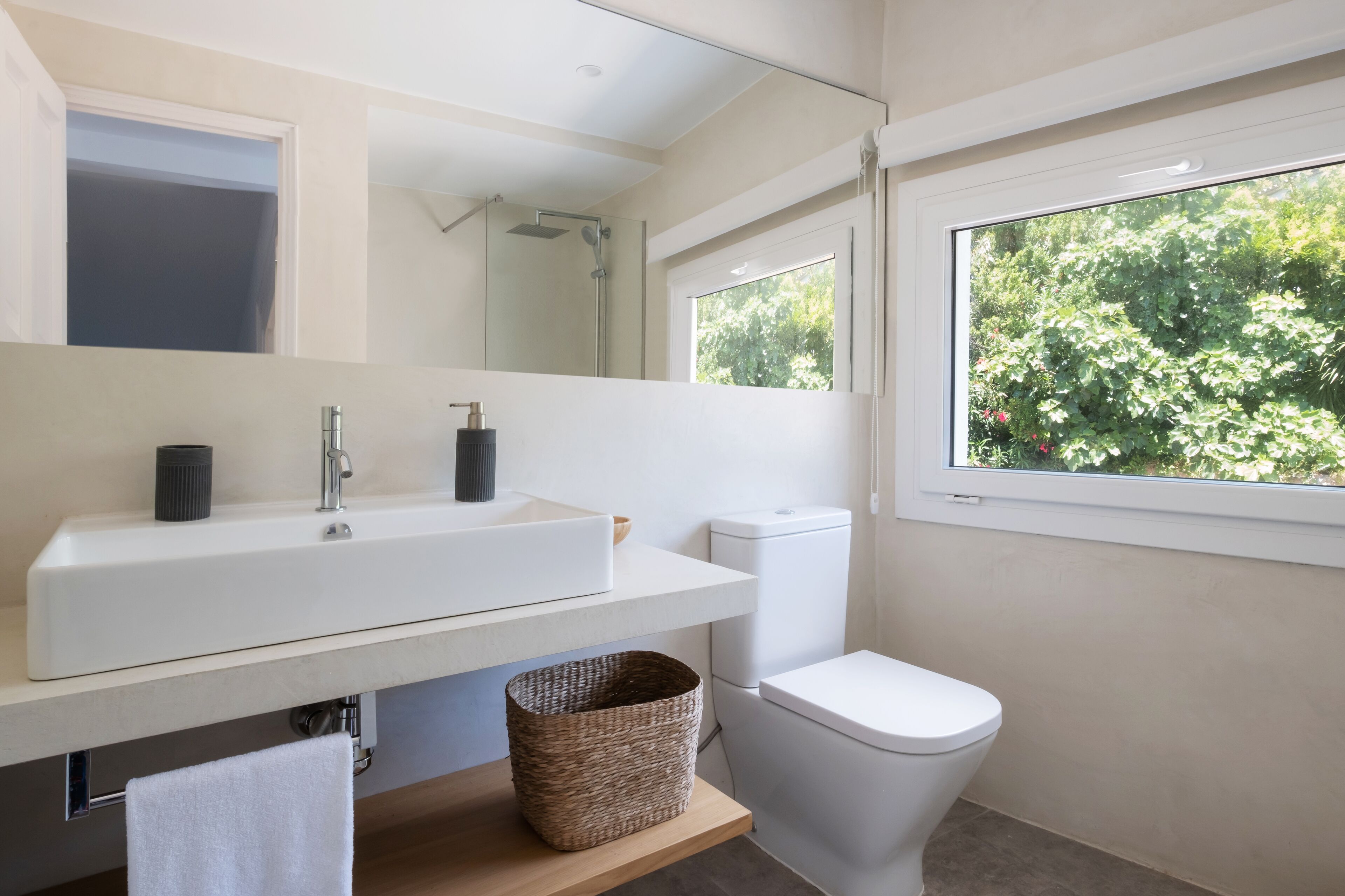A modern bathroom with a white sink, a mirror, a shower, a toilet, and a window with a view of greenery.