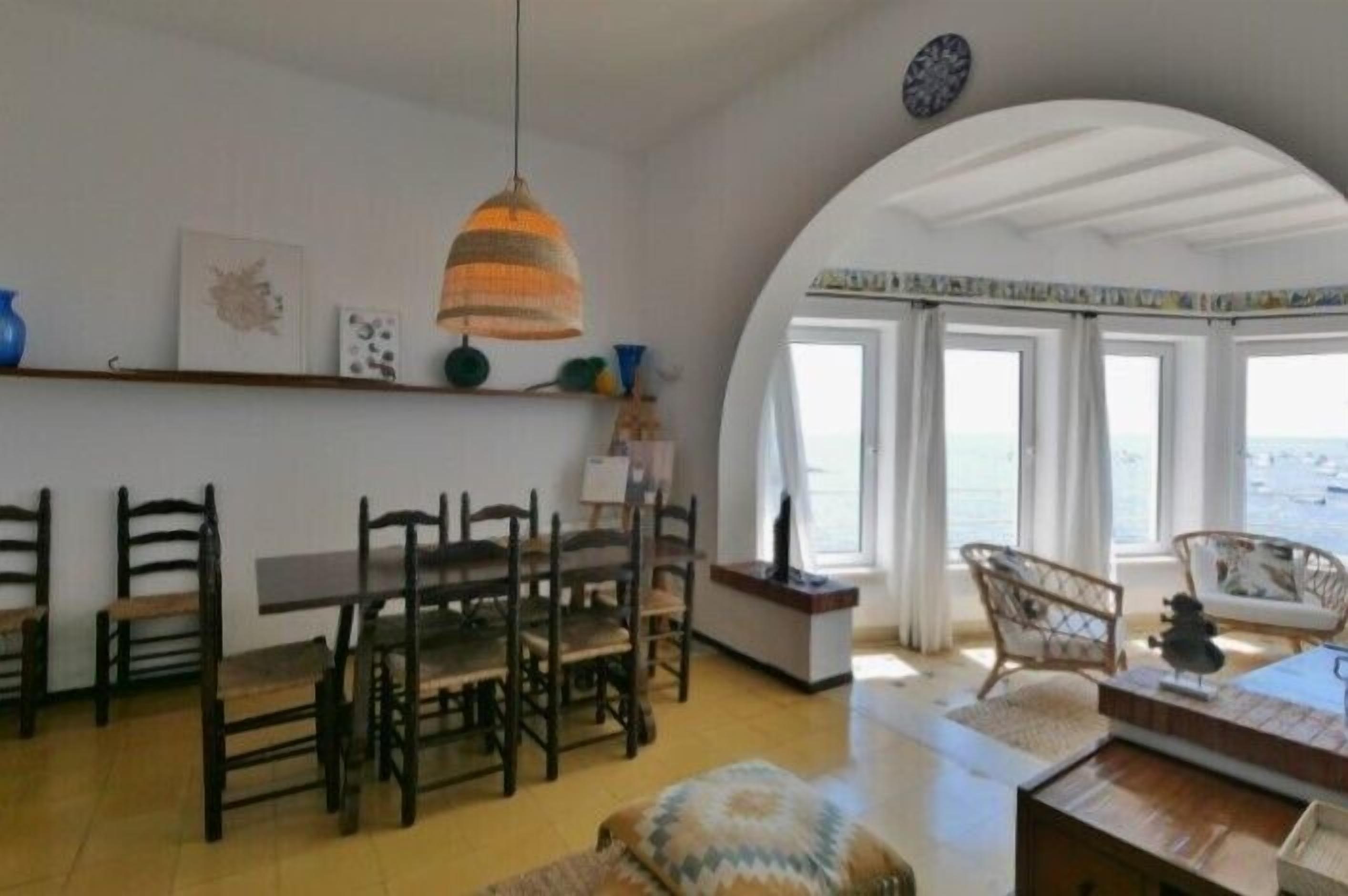Zona pranzo con tavolo e sedie in legno, una lampada a sospensione e vista mare attraverso ampie finestre.