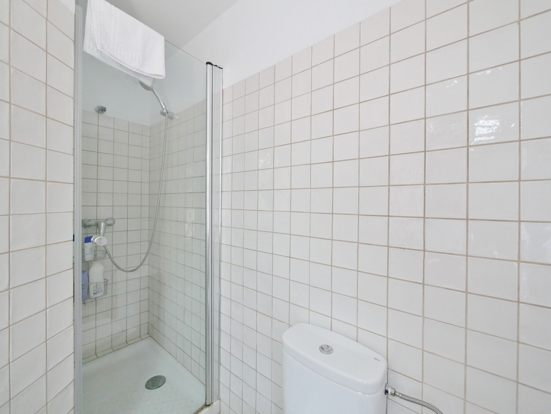 Une petite salle de bain propre, avec des carreaux blancs, une douche en verre et des toilettes blanches.