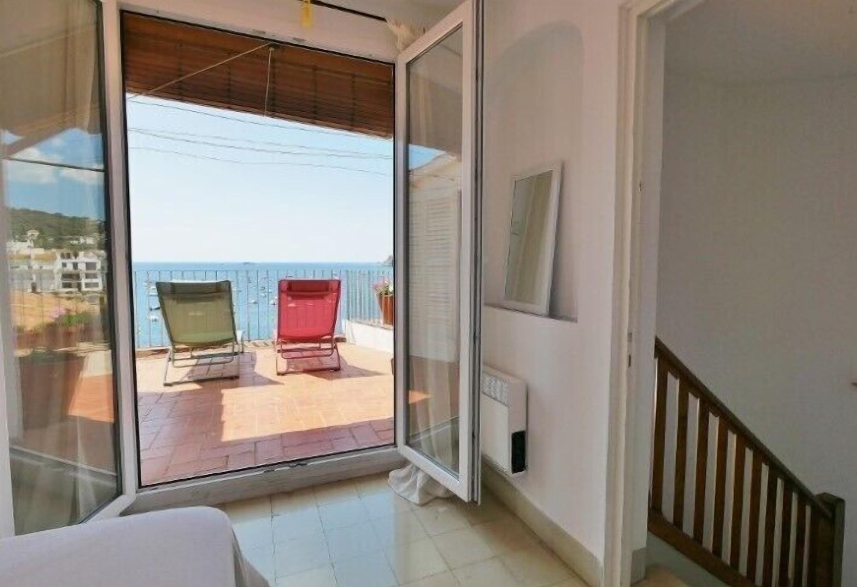 Una habitación con puerta de cristal corredera que da a un balcón con dos sillas y una mesa roja.