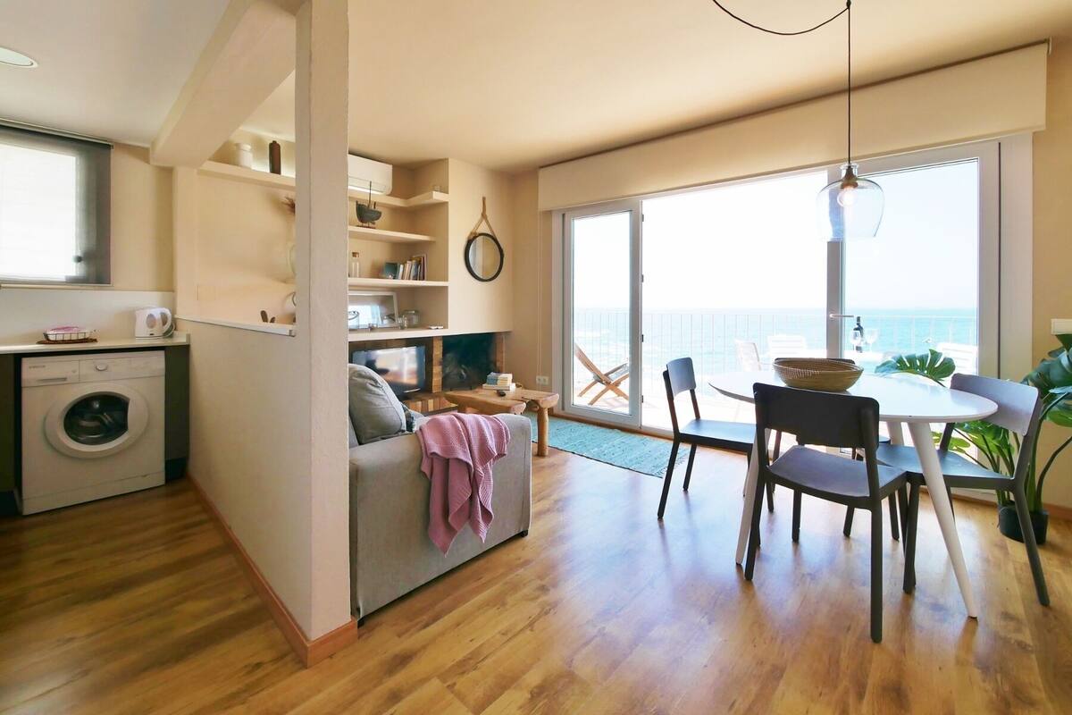 Un espacio habitable compacto con zona de comedor, lavadora y vista al mar a través de amplios ventanales.