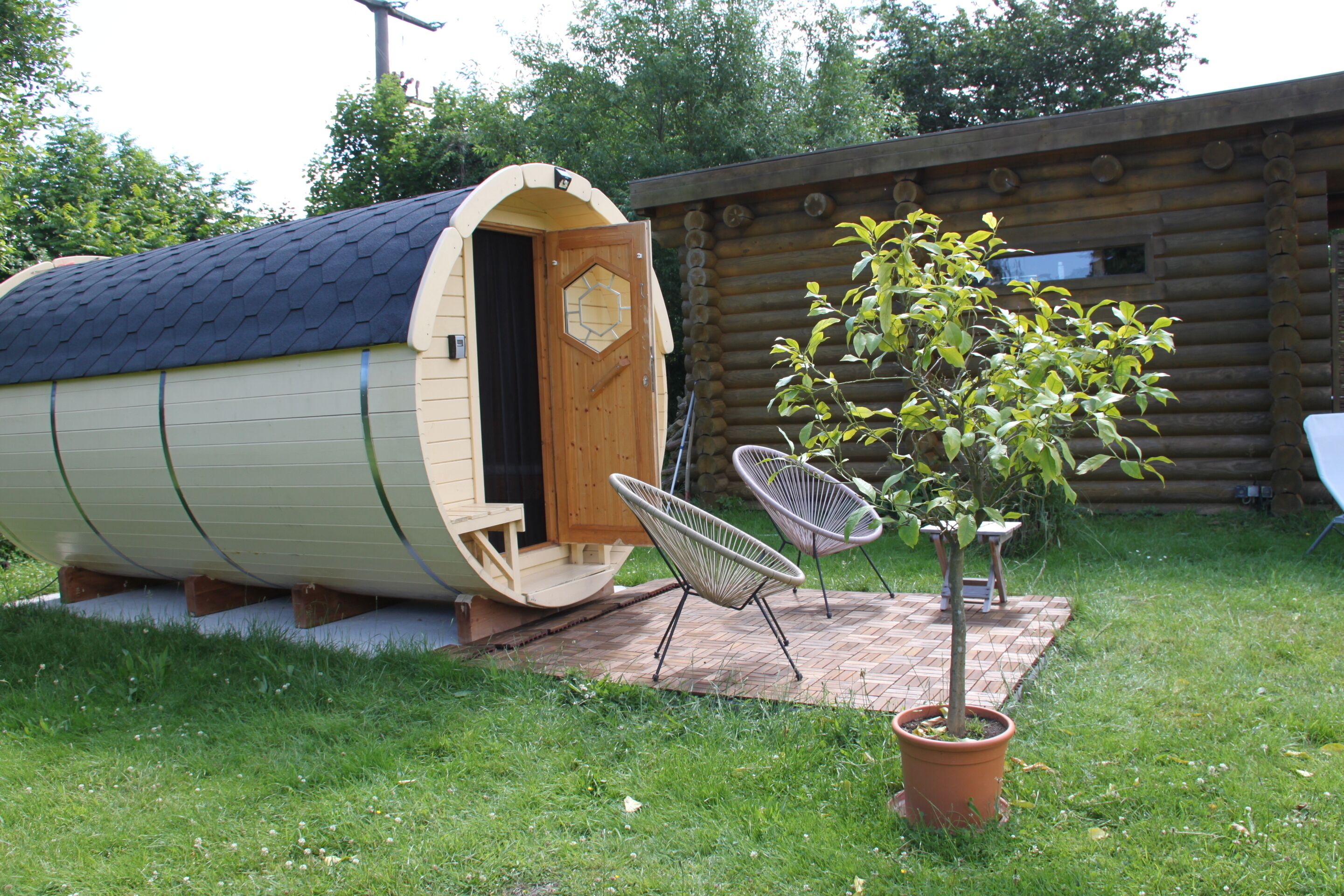 Una cabaña de troncos con una pequeña sauna adosada, dos sillas de mimbre y un árbol en maceta en el jardín.