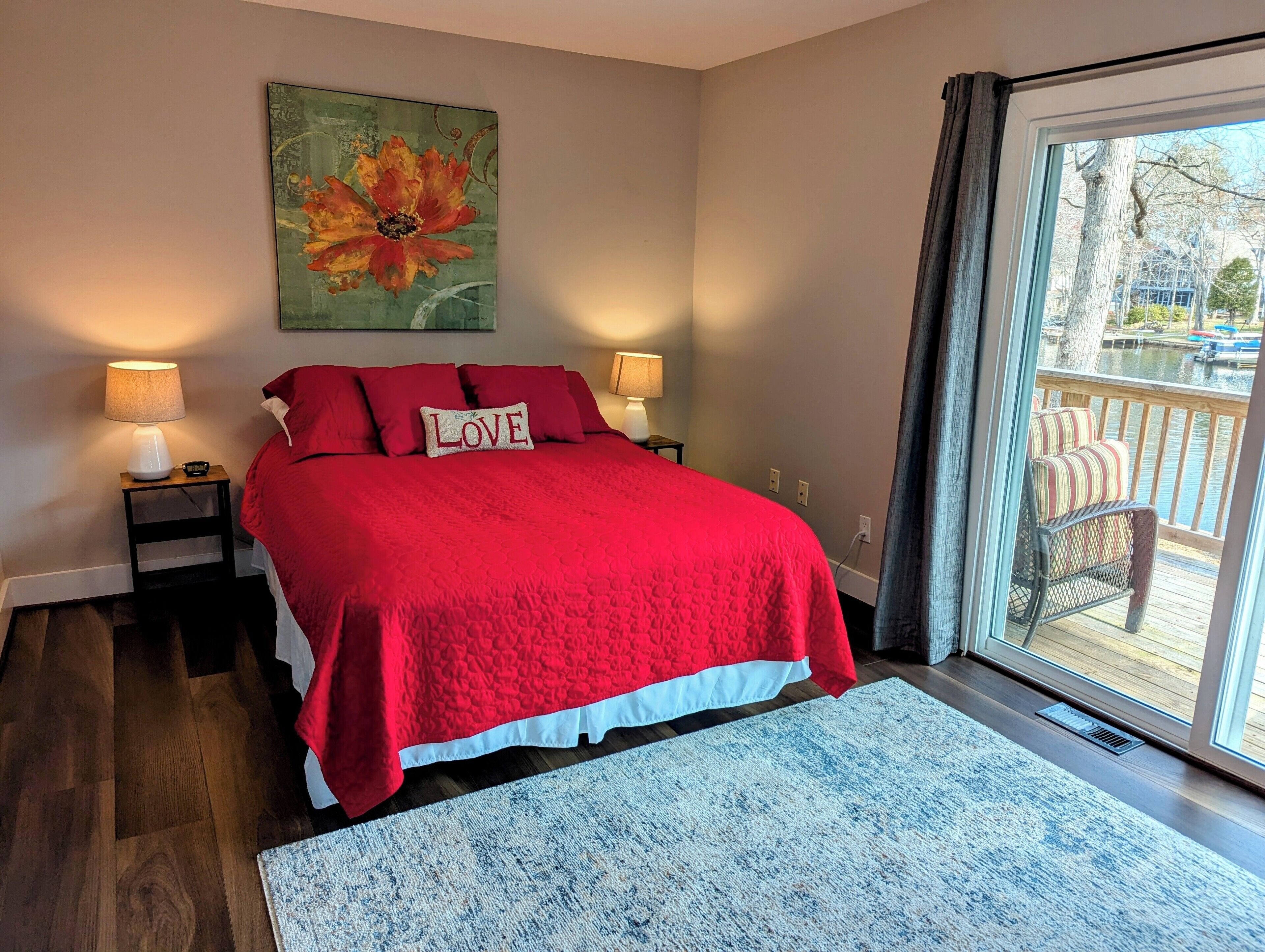 A bedroom with a large bed, two bedside lamps, a red throw blanket, a pillow with "LOVE" on it, a painting of a flower, and a view of a deck with outdoor furniture.