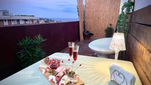 A table set with a charcuterie board, two glasses of red wine, and a white lamp on a balcony overlooking a cityscape.