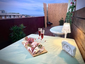 Une table dressée avec une planche de charcuterie, deux verres de vin rouge et une lampe blanche sur un balcon donnant sur un paysage urbain.