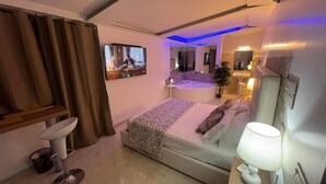 A modern hotel room with a bed, a bedside table with a lamp, a TV, and a bathroom visible through a glass door.